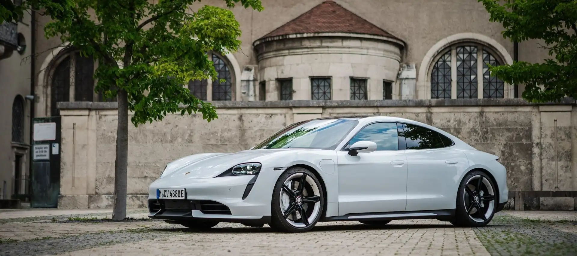 Our white Porsche Taycan seen from the side. It's located right under a tree in front of a museum.